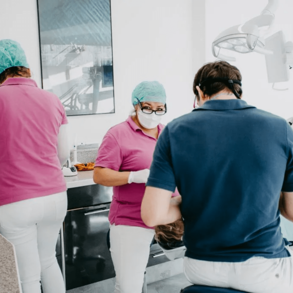 Behandlungssituation in der Zahnarztpraxis: Drei Mitarbeitende mit Schutzkleidung, Patient auf Behandlungsstuhl.