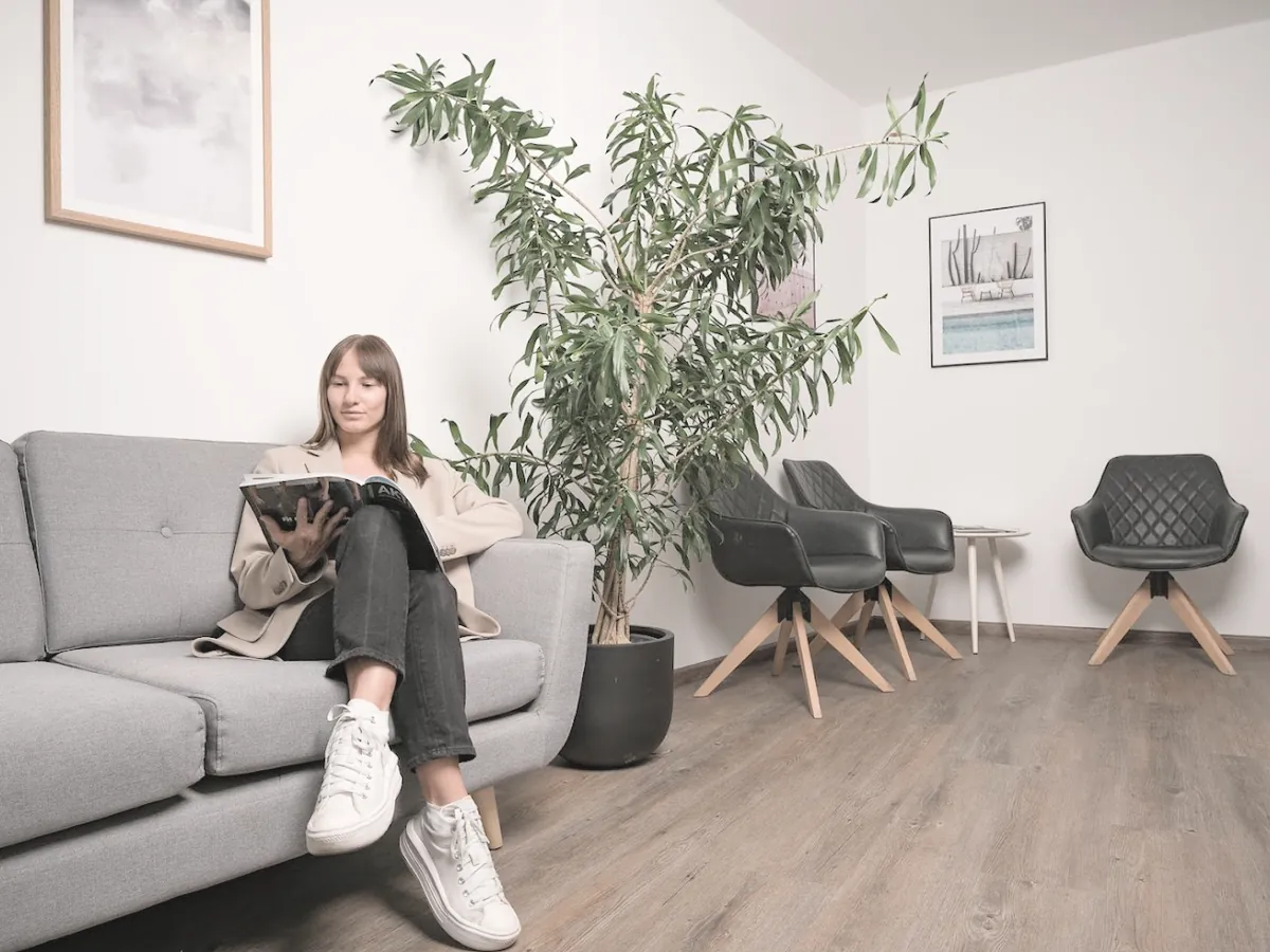 zahnarztpraxis-innsbruck-wartezimmer Wartezimmer der Zahnarztpraxis MeinZahnarzt in Innsbruck mit grauem Sofa, großer Grünpflanze und Sesseln, eine Patientin liest eine Zeitschrift